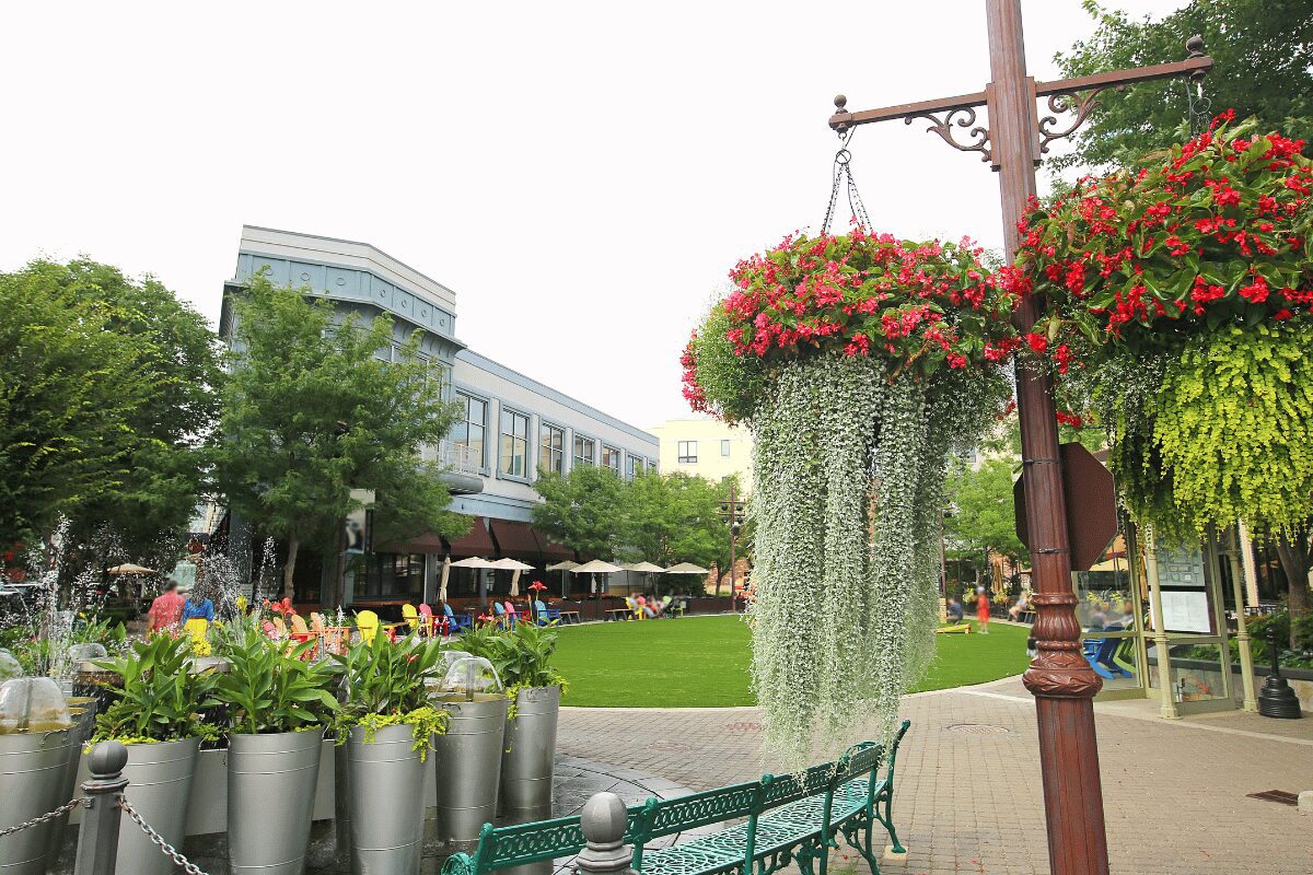 Outdoor shopping plaza with storefronts, fountains, hanging flower baskets and landscaped seating areas.