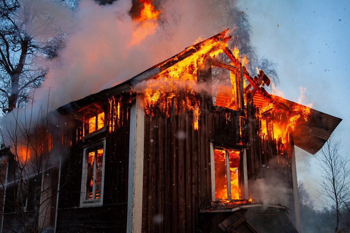 Two-story wood-sided building engulfed in flames, with heavy smoke and fire spreading through the roof and upper walls.