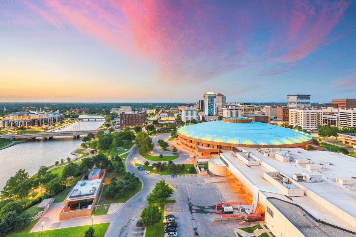 Aerial view of downtown Wichita, Kansas, at sunset with a circular arena, commercial buildings and riverfront bridges.